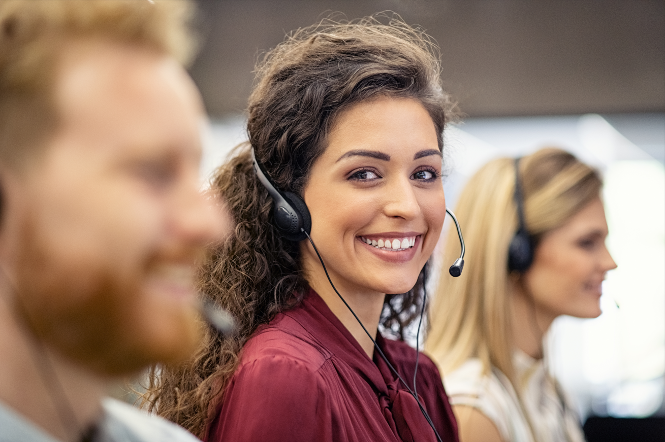 Female support center employee smiling at the camera while wearing a headset and providing assistance between two support center employees who are out of focus.