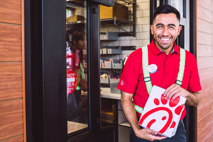 Closeup of a Hispanic male Chick-fil-A® Team Member smiling, holding a to-go bag of food outside of the drive-through window.