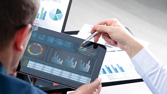 Over-the-shoulder closeup of a business analyst’s forearm and hand using a pointer to show circle and bar graphs on a tablet held by a client