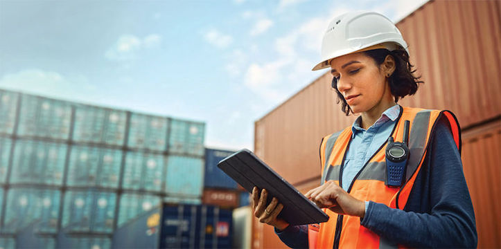Hispanic woman in a hardhat and orange safety vest working on a tablet in a cargo container yard.