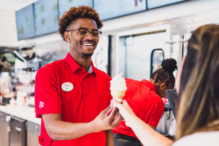 A black Chick-fil-A® Team Member smiles while serving a soft serve ice cream on a cone to a customer.