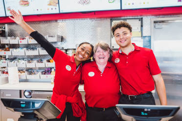 Two young black Chick-fil-A® Team Members, one male and one female, standing in a Chick-fil-A® kitchen on either side of an older white female Team Member. They are all smiling with their arms around each other.