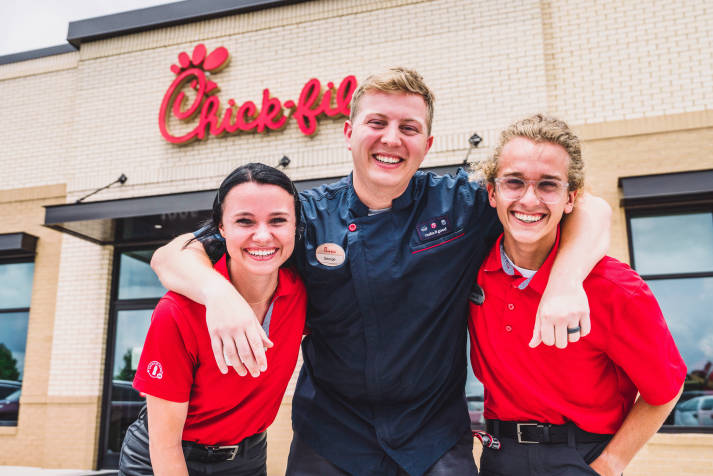 Closeup of a Chick-fil-A® manager standing between two Team Members. They are all smiling in front of a Chick-fil-A® restaurant with the sign visible in the background.