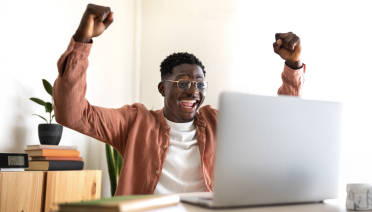Young black man sitting at a desk in front of a laptop computer smiling and cheering with his hands in the air.