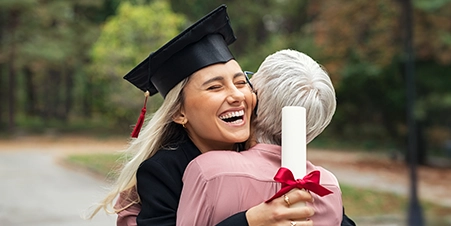 White female college graduate wearing a cap and gown, smiling and holding her degree while hugging her grandmother