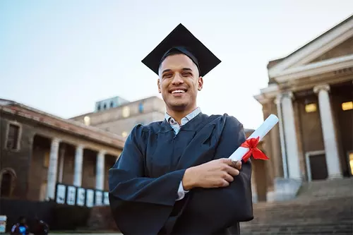Hispanic male in a cap and gown holding a degree on a college campus