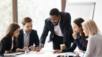 A project manager standing at a table pointing to a group of papers with graphs and other data. He is surrounded by coworkers sitting down