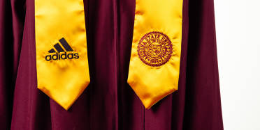 Closeup of the torso of an Arizona State University graduate wearing a graduation gown with and an untied necktie with the Adidas logo on one side and the Arizona State University logo on the other