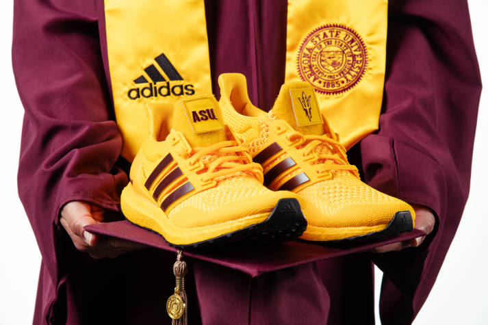 Closeup of the torso of an Arizona State University graduate wearing a graduation gown and holding a pair of Arizona State University colored Adidas shoes.