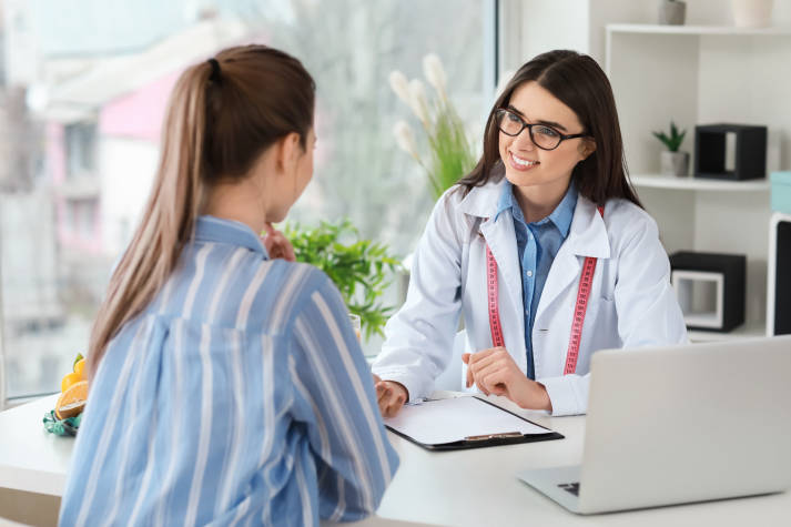 Female Intermountain caregiver wearing a lab coat sits at a desk across from a patient.