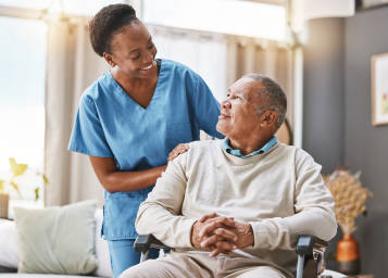 Black female Intermountain caregiver wearing blue scrubs stands smiling over a sitting older black male patient who is smiling back at her.