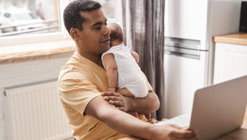 Black adult learner holding their baby while doing schoolwork on a laptop at home.