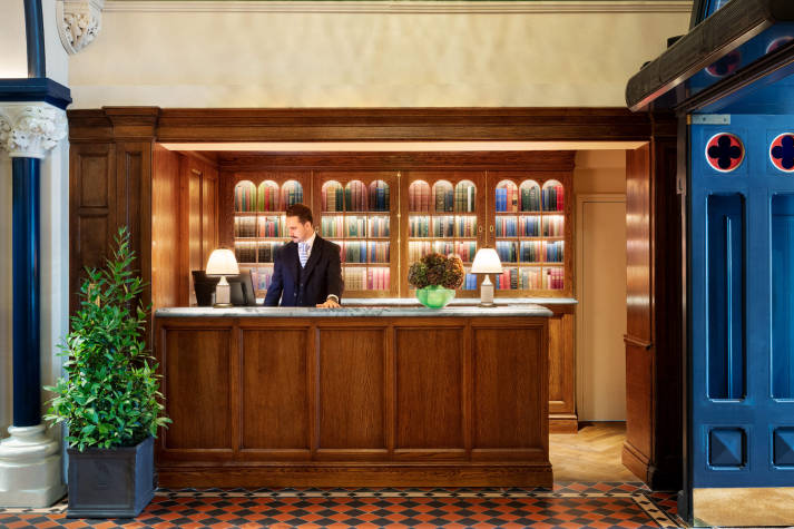 Male Graduate Hotel associate in a dark suit standing behind the front desk of a Graduate Hotel. 