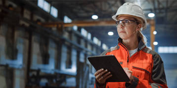White female industrial engineer wearing a hardhat and safety glasses, working on a tablet in a warehouse