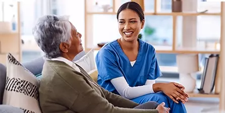 Asian female Intermountain caregiver wearing blue scrubs sits on a couch laughing with a patient.