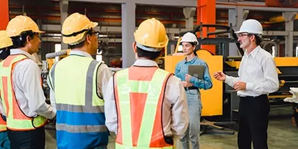 Male and female supervisor, wearing hard hats in a warehouse speaking to a group of employees seen from behind wearing hardhats and safety vests