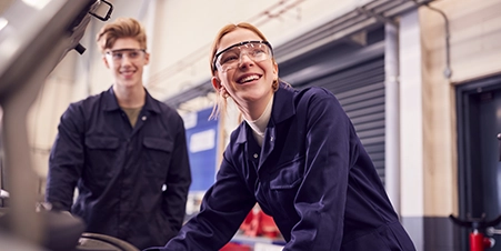 White female wearing mechanic’s overalls and safety glasses, looking under the hood of a car. Her white male coworker is standing next to her.