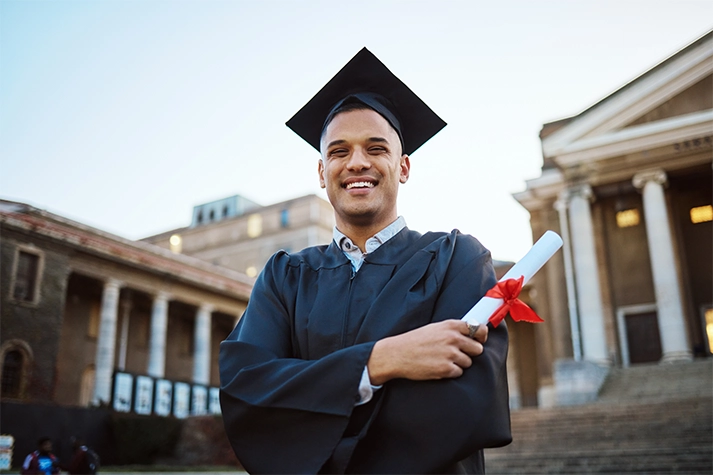 Hispanic male in a cap and gown holding a degree on a college campus.