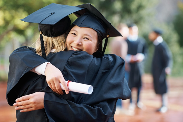 Asian female graduate in a cap and gown, holding a rolled-up diploma, hugs another graduate whose back is to the camera