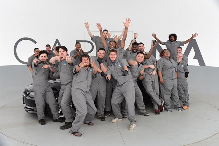 Large group of Carvana employees wearing gray mechanic suits, leaning against a car, smiling and posing with thumbs up in a studio space with “Carvana” displayed behind them