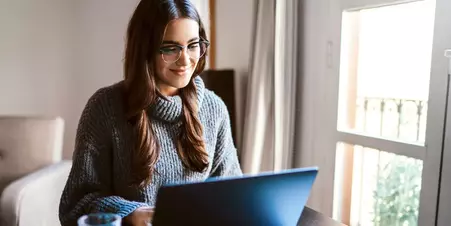 Woman in a grey turtleneck sweater and glasses sitting at a home office looking at a laptop and smiling.