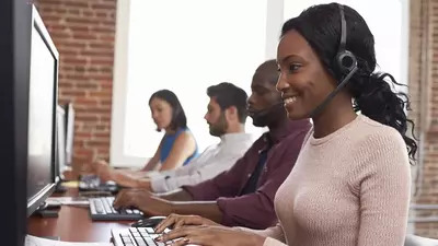 Black female support center employee wearing a headset and working at a computer while providing assistance.