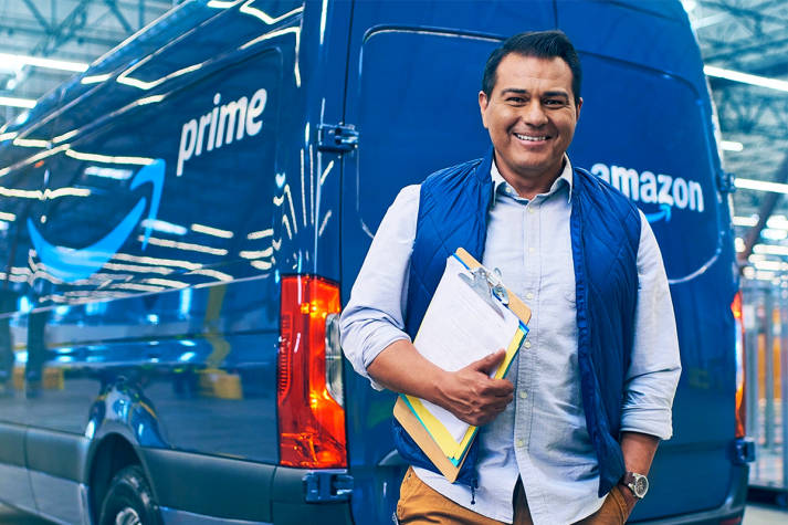 Hispanic DSP owner smiling, holding a clipboard and standing in front of an Amazon delivery van parked inside a distribution center.