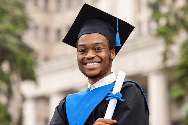 Closeup of a black male graduate in a cap and gown holding a rolled up diploma to his left shoulder while smiling down at the camera.