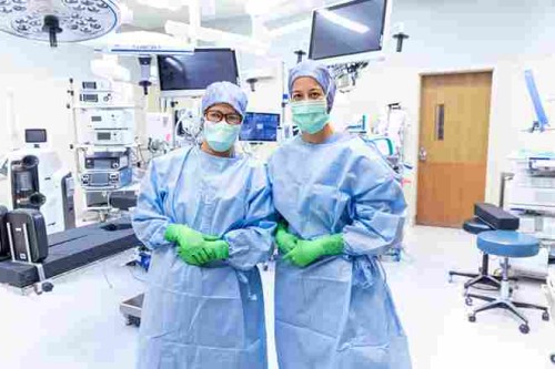 Two white female surgical technicians wearing surgical gowns, hair coverings, gloves and face masks in an operating room