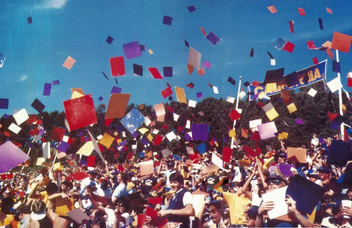 Vintage photo of a large group of people throwing multi-colored graduation caps under a blue banner that says “California”