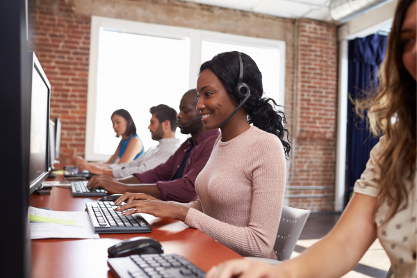 Black female support center employee wearing a headset and working at a computer while providing assistance