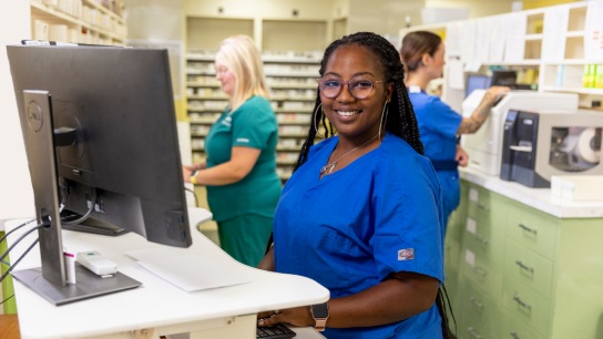 Black female pharmacy technician working at a desktop computer in a pharmacy and two white female technicians working in the background