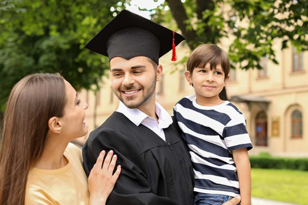White male wearing a cap and gown on a college campus, smiling at his wife and carrying his young son