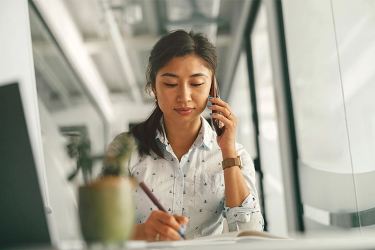 Asian woman sitting at a desk, talking on a cell phone and taking notes
