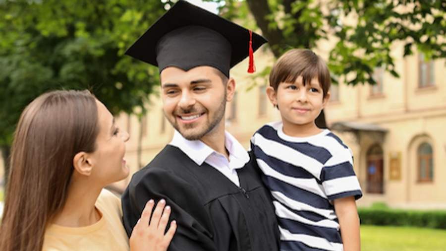 White male in a cap and gown at his graduation ceremony with his wife and young son