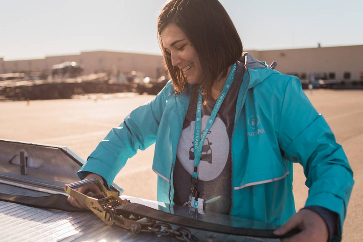 White female Carvana employee wearing a Carvana shirt, lanyard and jacket adjusting a strap on a hauler in a parking lot