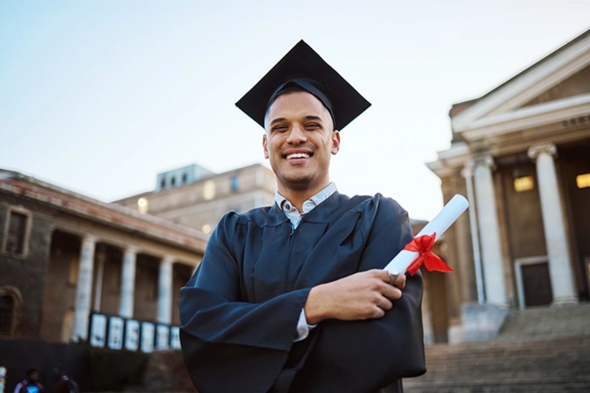 Hispanic male in a cap and gown holding a degree on a college campus
