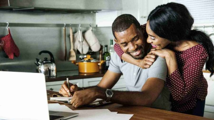 Black man smiling, sitting at a kitchen counter with a laptop open in front of him while he takes notes in a notebook, his Black wife hugging him from behind