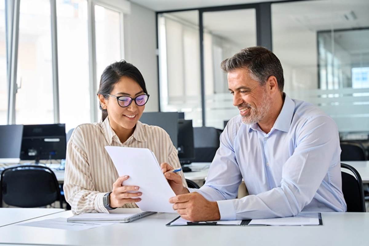 Asian female and Hispanic male sitting at a conference table in an open office, referring to a document