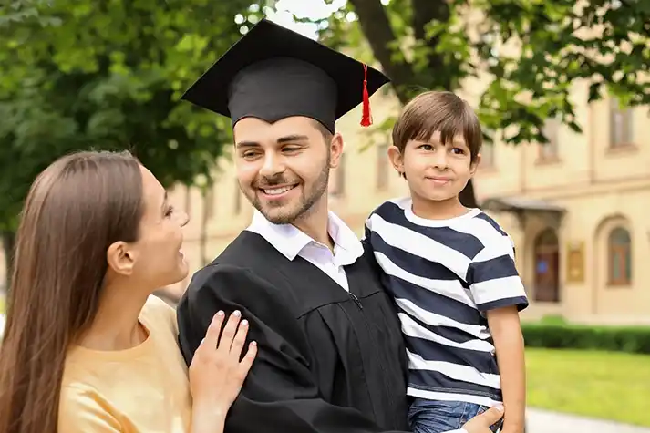 White male wearing a cap and gown on a college campus, smiling at his wife and carrying his young son