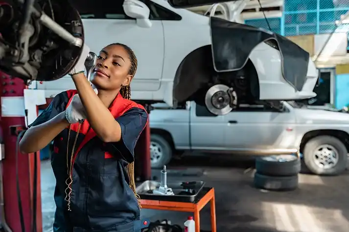 Black female wearing mechanic’s overalls working on a car in an auto shop.