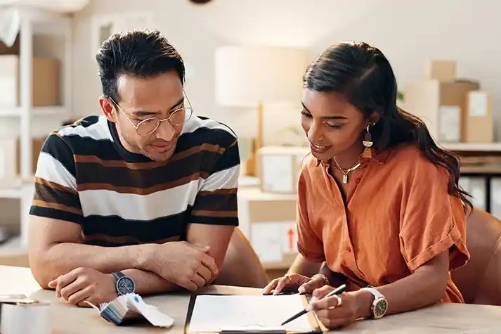 A man and woman sit at a table and look at information on a clipboard.