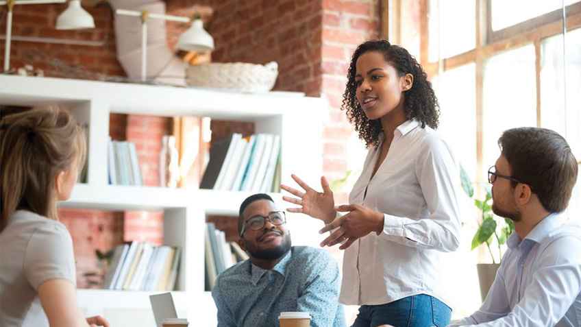 Black female in a conference room standing up and speaking to a team of coworkers seated around her