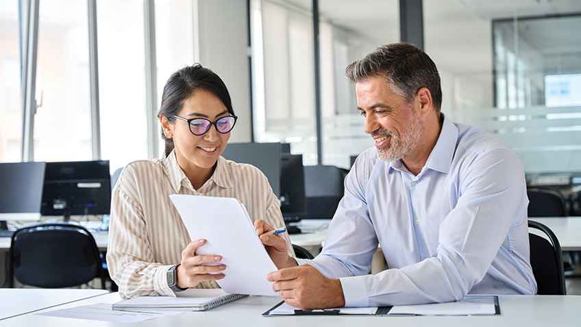 An Asian woman and a white man with a salt and pepper beard hold a piece of paper together while he points to something on the paper.