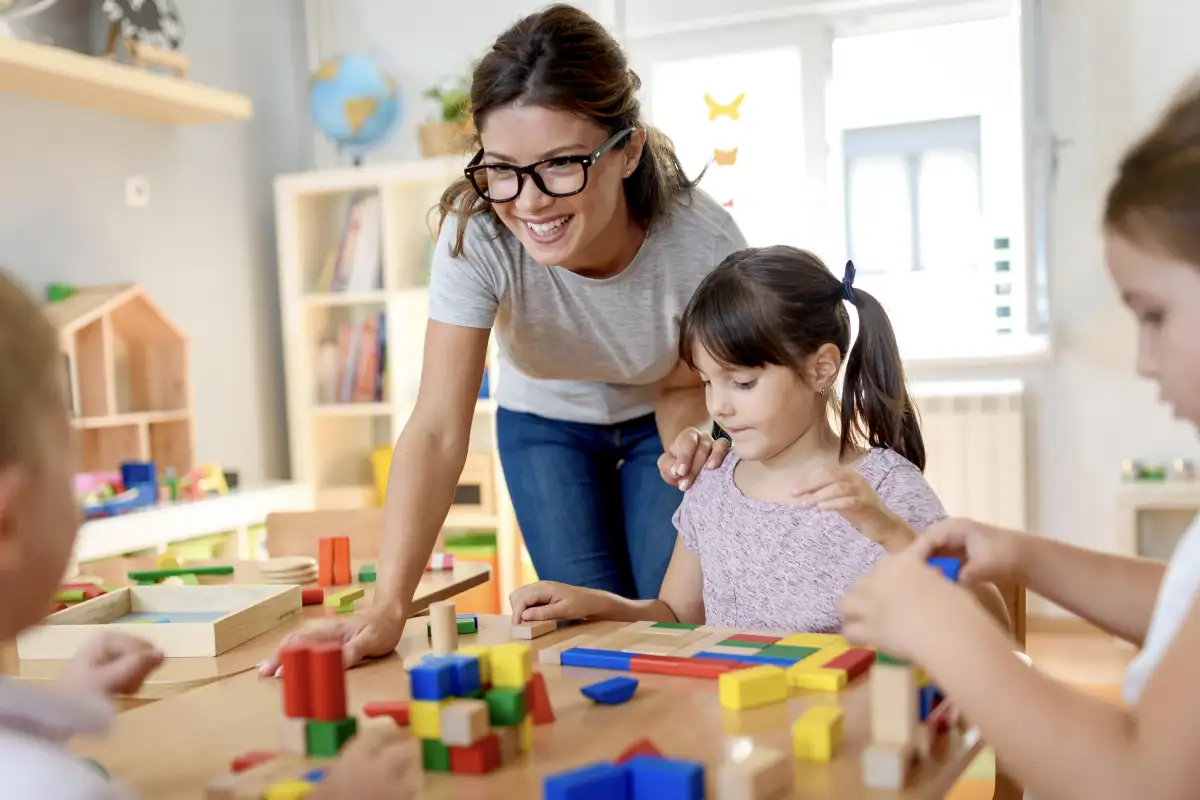Female early child educator in a classroom leaning over a female student playing with blocks while two other students can be seen out of focus on the edges of the image