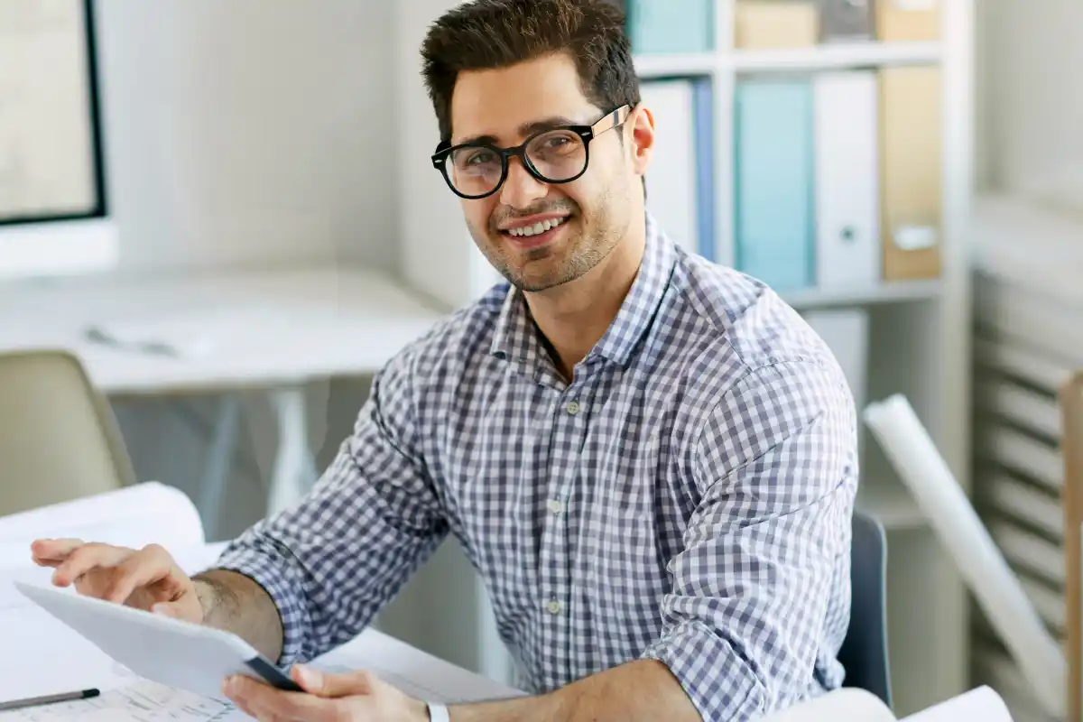 Smiling man in glasses and a checkered shirt sitting at a desk, holding a tablet