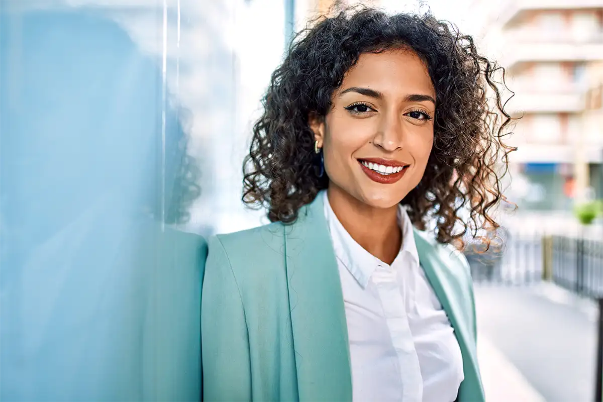 Closeup of a Latina female smiling at the camera in a teal blazer and white dress shirt standing outside leaning against a wall
