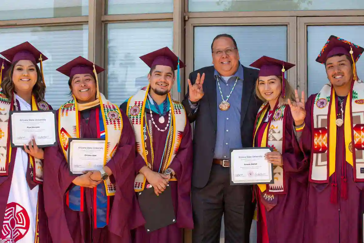 Five Gila River Indian Community graduates, three female and two male, stand in crimson ASU caps, gowns, and sashes with traditional Gila River colors and patterns alongside a male Gila River Indian Community leader flashing the ASU devil horn symbol with one of the graduates.