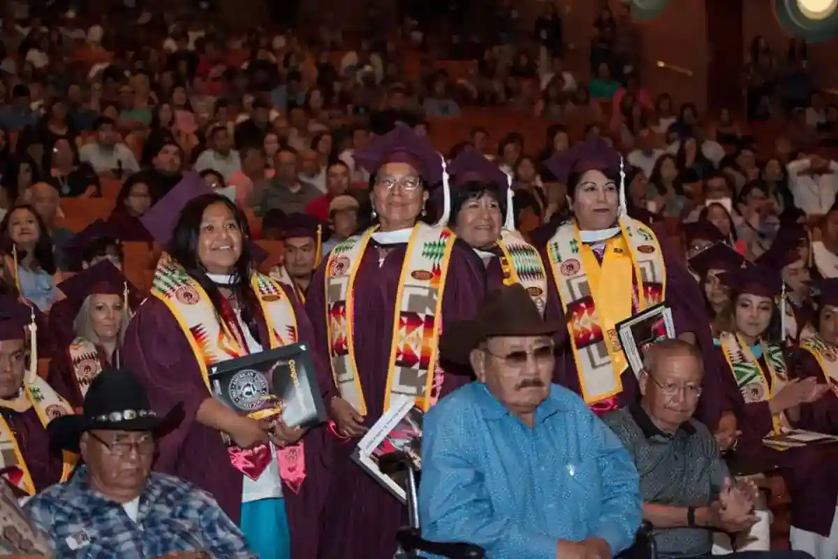 Four female graduates from the Gila River Indian Community stand in crimson ASU caps and gowns, wearing sashes with traditional Gila River colors and patterns, in a crowded auditorium filled with fellow graduates and their families.