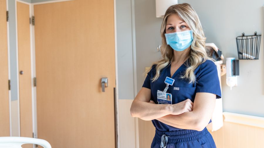 Rochester Regional Health team member in a surgical mask and blue scrubs standing in an exam room smiling at the camera